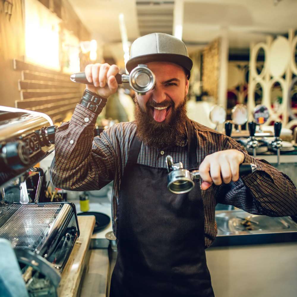 Funny young bearded man covering one eye with cezve and showing tongue on camera. He stands in kitchen at coffee machine.