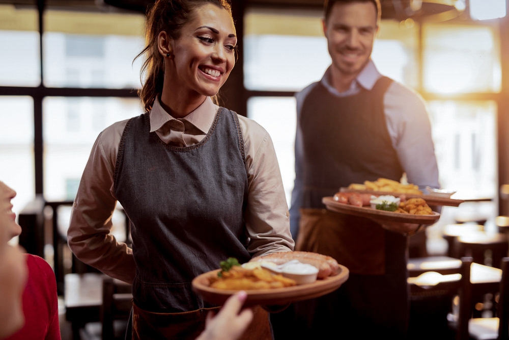 two waiters serving lunch and brining food to their gusts in a tavern. Focus is on happy waitress.