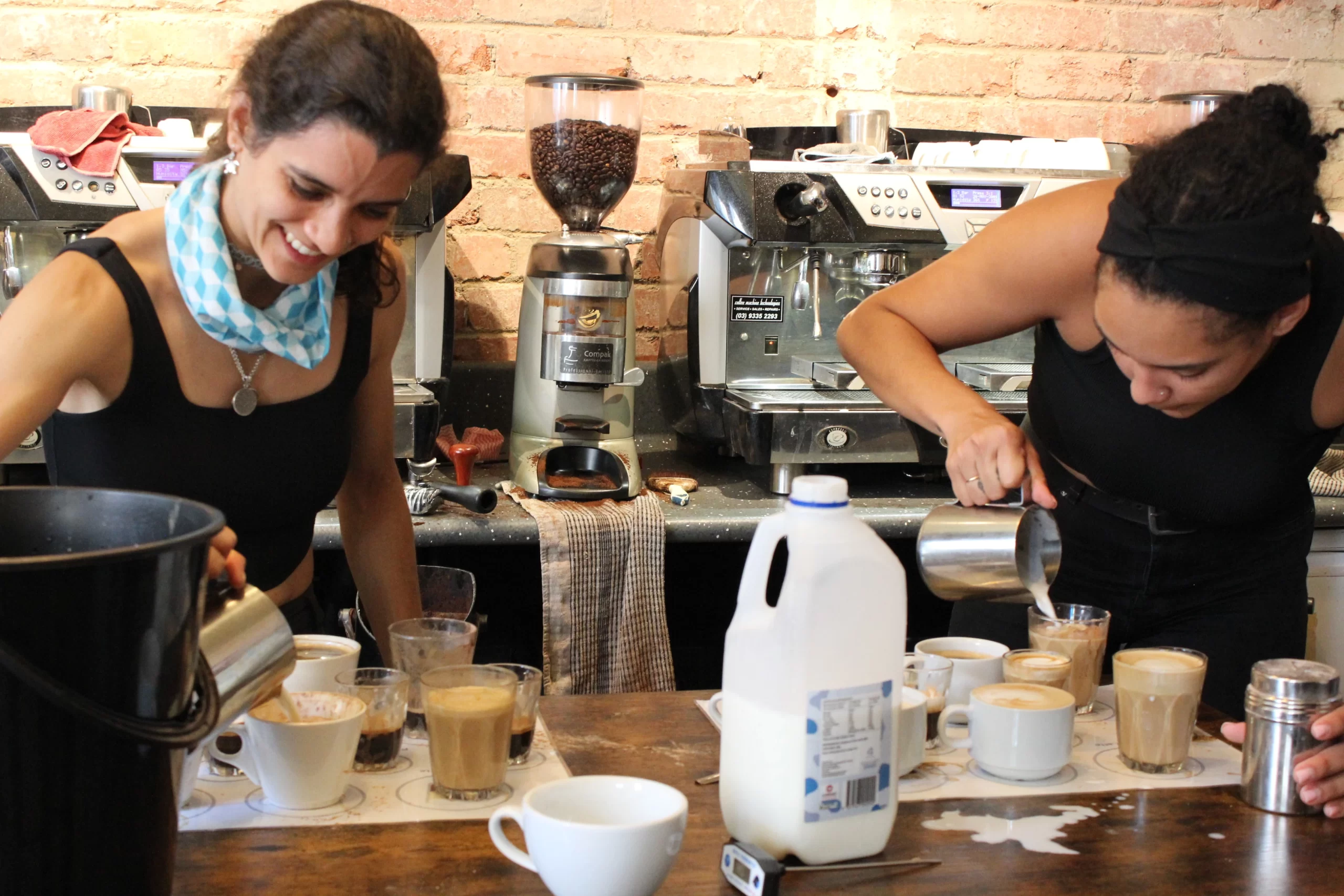 Two baristas preparing multiple coffee drinks, pouring steamed milk into cups behind a café counter with an espresso machine and grinder.